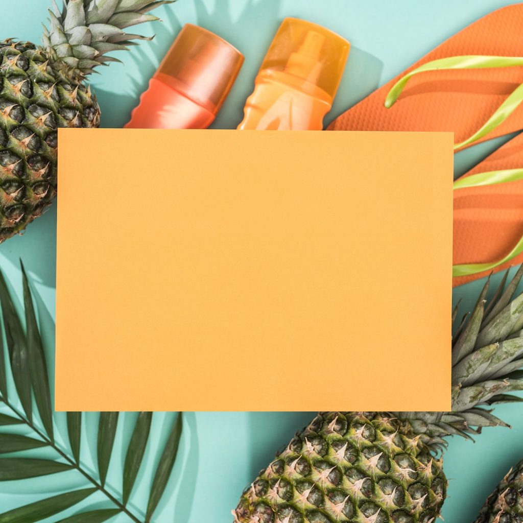 top view of pineapples, tropical leaf, sunscreens, orange flip flops and empty card on turquoise